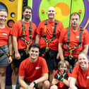 Image shows the seven members of the Calibre Climb Team standing in front of a climbing wall wearing red Calibre Audio T-shirts and climbing harnesses. Photo from Andy Scott Photohello